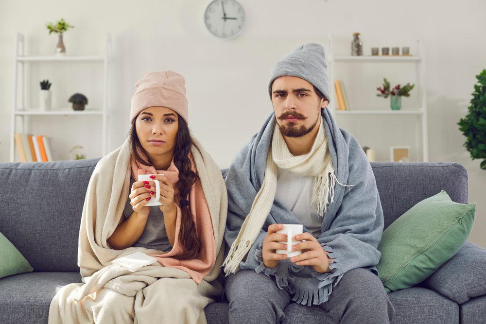 Dissatisfied,Young,Couple,Having,Problem,With,Central,Heating,,Sitting,On ;ietminderung - Paar mit Heißgetränken auf Couch, kalt wirkend.