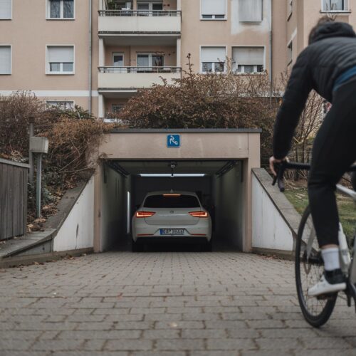PKW fährt langsam aus Garage während Radfahrer auf Gehweg nähert