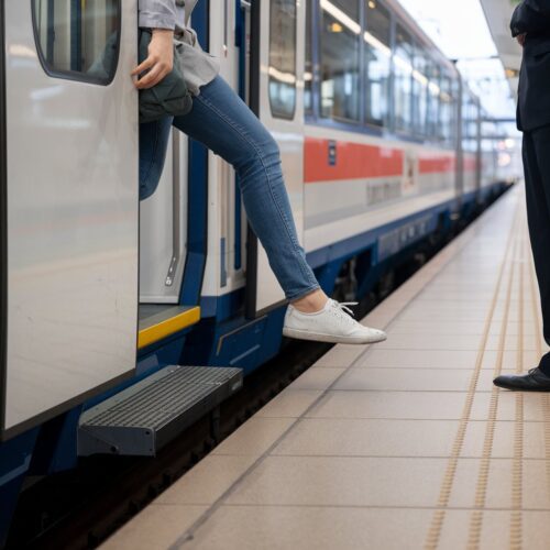 Frau mit einem Fuß auf Zugstufen, der andere Fuß nahe dem Bahnsteig, große Lücke zwischen Zug und Plattform.