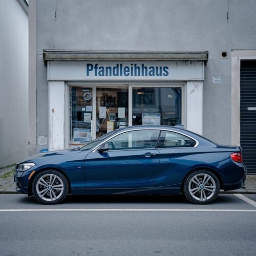 Dunkelblauer BMW 120d vor unscheinbarem Pfandhaus mit einfachem Schild.