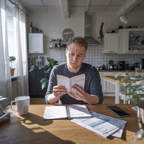 Mann in Wohnzimmer notiert PIN in Notizbuch, wirkt besorgt. Schreibtisch mit Alltagsgegenständen.