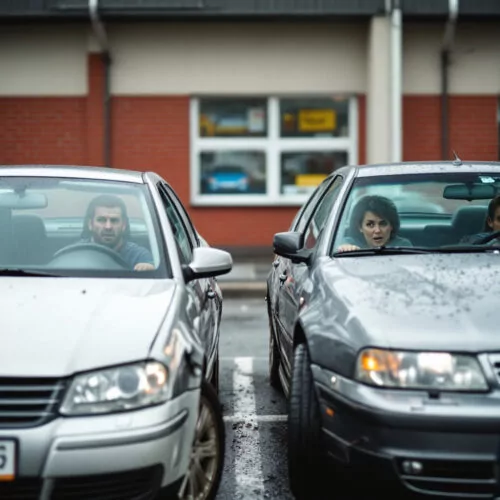 Zwei Autos kollidieren im Parkplatz eines Supermarkts; Fahrer schauen überrascht.