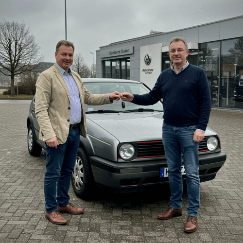 Autoübergabe vor einem Autohaus: Verkäufer übergibt Schlüssel an Käufer, VW Golf auf nassen Pflastersteinen.