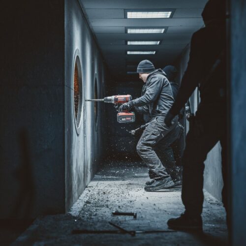 Burglars drilling into a bank vault in a dimly lit corridor, with dust in the air and tools scattered on the floor.